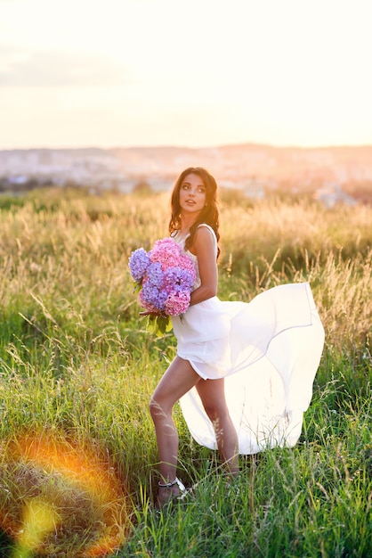 Zmysłowa śliczna dziewczyna w lekkiej białej letniej sukience z bukietem delikatnych kolorowych kwiatów chodzi po trawniku o zachodzie słońca.