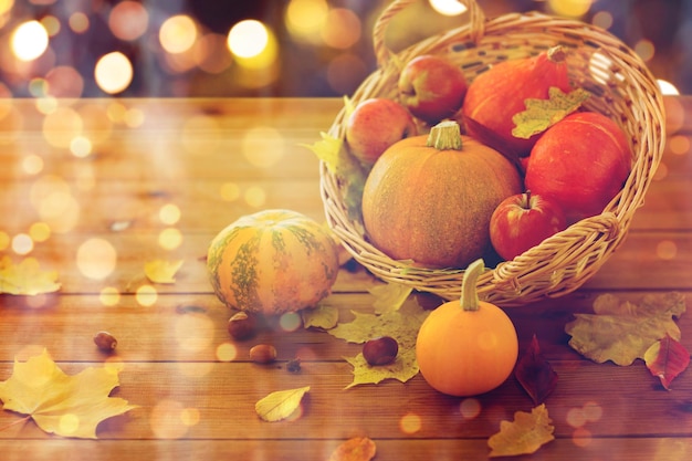 żniwa, sezon, halloween i jesień koncepcja - zbliżenie dyni w wiklinowym koszu z liśćmi na drewnianym stole nad świątecznymi światłami