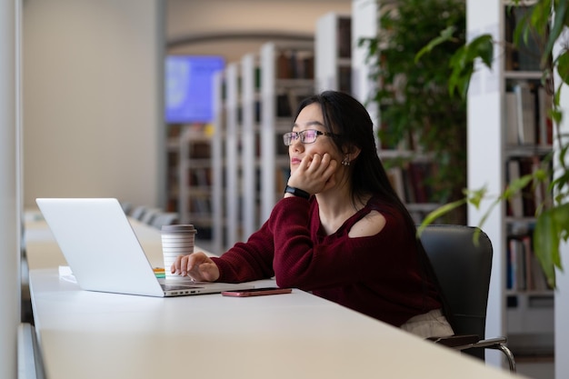 Znudzona azjatycka studentka patrzy na laptopa, siedzi przy biurku w bibliotece, nie koncentruje się na lekcji online