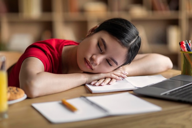 Znudzona zmęczona młoda koreańska studentka śpi na biurku z laptopem we wnętrzu pokoju