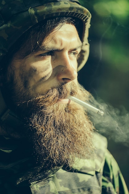 Żołnierz z zawiązaną i brudną brodatą twarzą w hełmie wojskowym i amunicją palący papierosa