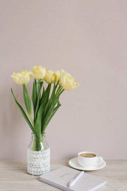 Żółte tulipany w wazonie na stole filiżanka kawy i pamiętnik z długopisem szarobeżowa ściana