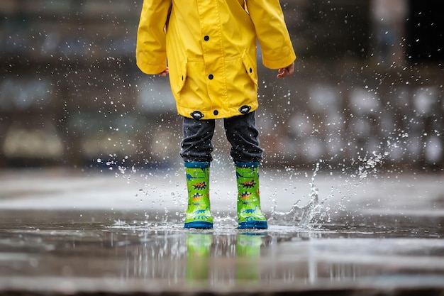 Żółty płaszcz przeciwdeszczowy dziecka i zielone buty wpadają do kałuży wody. Jesienna deszczowa pogoda.