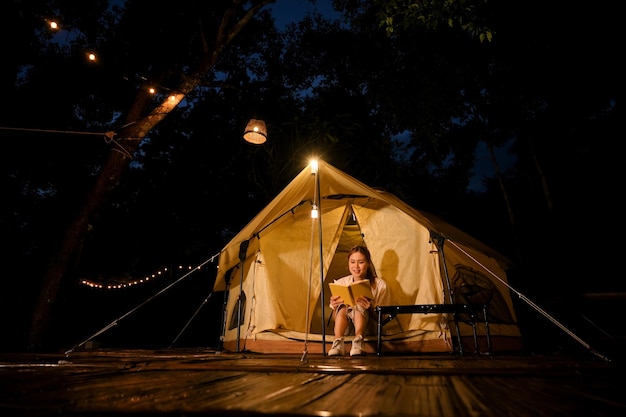 Zrelaksowana Azjatka siedząca przed swoim namiotem i czytająca książkę w nocy