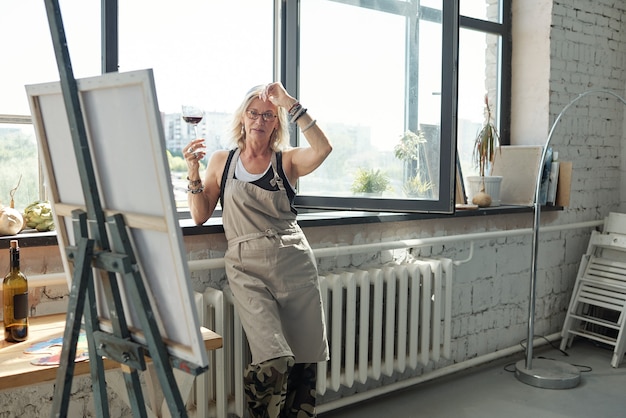 Zrelaksowana dojrzała artystka w fartuchu stojąca przy oknie i pijąca wino w pracowni artystycznej