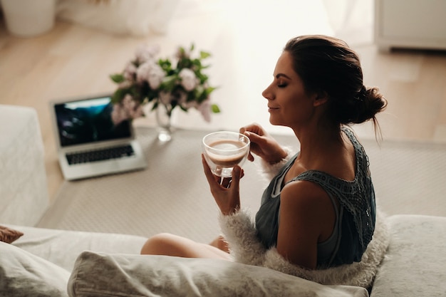 Zrelaksowana dziewczyna w domu pije kawę. Wewnętrzny spokój. Dziewczyna siedzi wygodnie na kanapie i pije kawę
