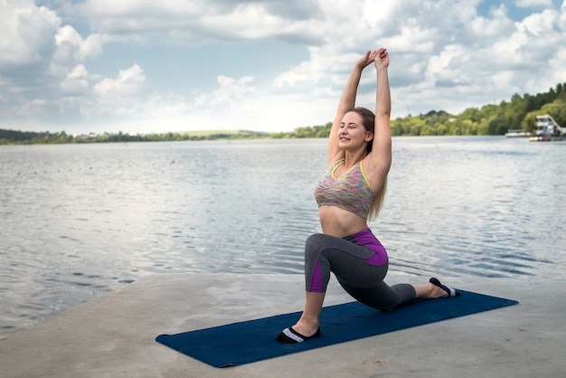 Zrelaksowana dziewczyna w stroju sportowym wykonująca ćwiczenia rozciągające, medytacyjne, oddechowe na macie do jogi w pobliżu jeziora