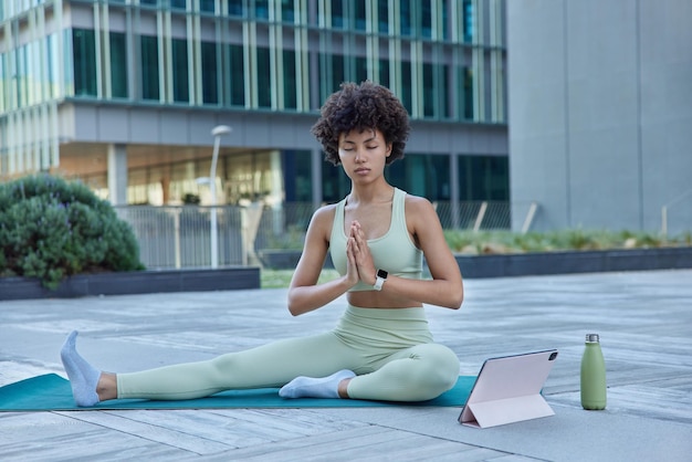 Zrelaksowana, kręcona kobieta ćwiczy jogę zen, ubrana w sportowe pozy na macie fitness z cyfrowym tabletem i butelką wody, cieszy się poranną medytacją w miejskim otoczeniu. Strzelanie do całego ciała