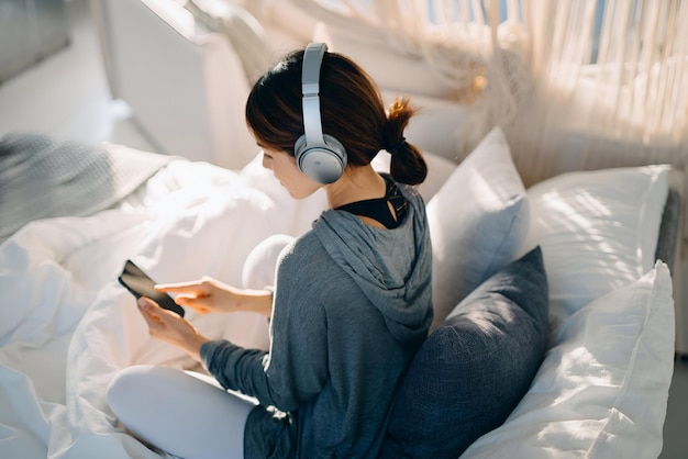 Zrelaksowana młoda Azjatka siedzi na łóżku, słucha muzyki przez słuchawki i surfuje po sieci za pomocą smartfona w domu