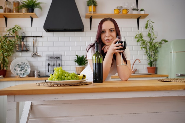 Zrelaksowana młoda kobieta pozuje stojąc przy kuchennym stole Dorosła brunetka odpoczywa z alkoholem i owocami w kuchni