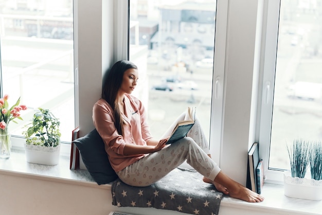 Zrelaksowana młoda kobieta w przytulnej piżamie czyta książkę, odpoczywając na parapecie w domu