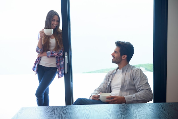 zrelaksowana młoda para pije pierwszą poranną kawę nad dużym jasnym oknem w nowoczesnym wnętrzu willi