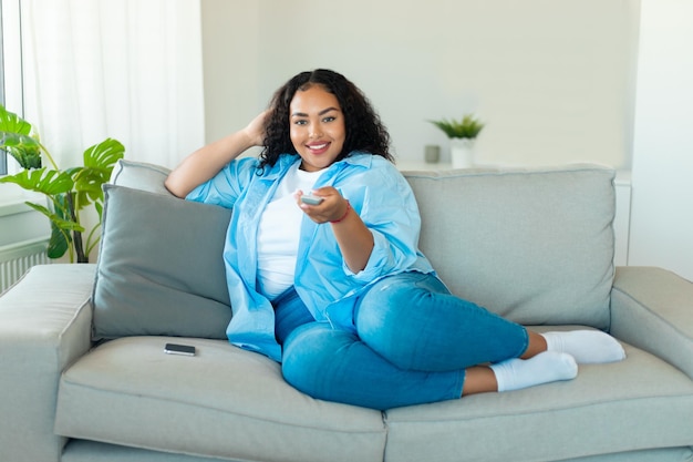 Zrelaksowana, pozytywna kobieta o czarnym ciele, przełączająca kanały telewizyjne z pilotem oglądającym telewizję na kanapie w domu