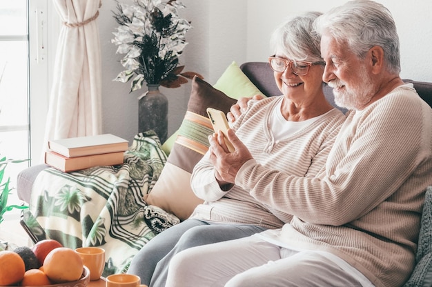 Zrelaksowana starsza para siedząca razem na domowej kanapie za pomocą telefonu komórkowego, ciesząca się technologią i społecznością