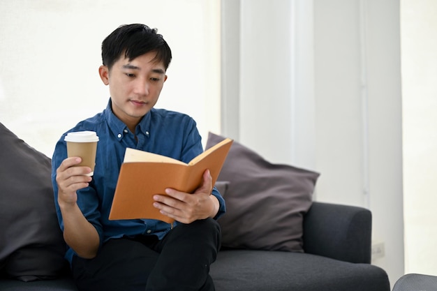 Zrelaksowany Azjata popijający kawę i czytający książkę na kanapie w salonie