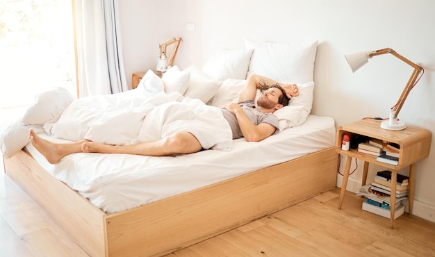 Zrelaksowany mężczyzna śpiący w łóżku w domu zmęczony człowiek śniący w sypialni i odpoczywający na wakacjach w domu Wyczerpany mężczyzna relaksujący się w hotelu rano wyglądający przytulnie i śpiący na miękkiej pościeli