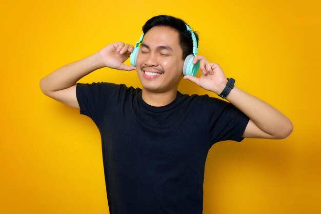 Zrelaksowany młody azjatycki człowiek w casualowej koszulce słuchający muzyki w słuchawkach na żółtym tle Ludzie emocje koncepcja stylu życia