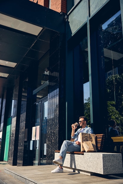 Zrelaksowany młody człowiek pijący aromatyczną kawę w pobliżu centrum handlowego i siedzący w pobliżu swoich toreb