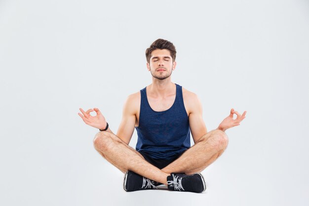 Zrelaksowany młody fitness człowiek z zamkniętymi oczami, siedzący w pozycji lotosu i medytujący na białym tle