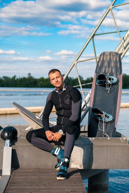 Zrelaksowany sportowy mężczyzna w piance, siedzący na molo z wakeboardem