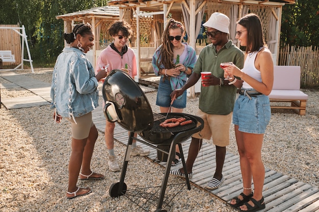 Zdjęcie zróżnicowana grupa przyjaciół smażąca hot dogi na grillu na imprezie na plaży, pijąca i rozmawiająca