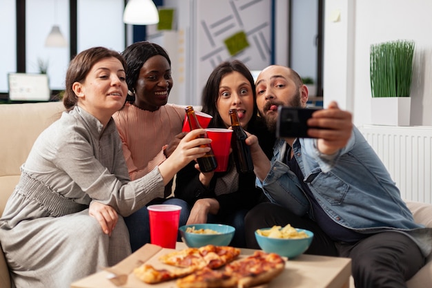 Zróżnicowana grupa współpracowników biorąca selfie na smartfonie po pracy na imprezie biurowej. Wesoli koledzy bawią się na uroczystym spotkaniu z butelkami z chipsami do pizzy i kubkami piwnego alkoholu