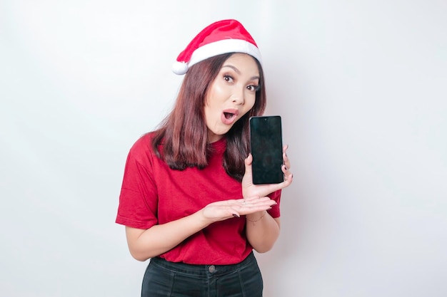 Zszokowana Azjatycka kobieta Świętego Mikołaja pokazuje swój telefon odizolowany przez białe tło Boże Narodzenie koncepcji
