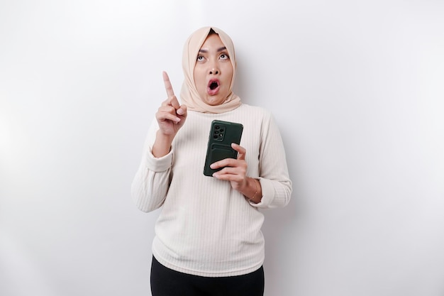 Zszokowana azjatycka muzułmanka nosząca hidżab, wskazująca na miejsce na kopię, trzymając telefon odizolowany białym tłem