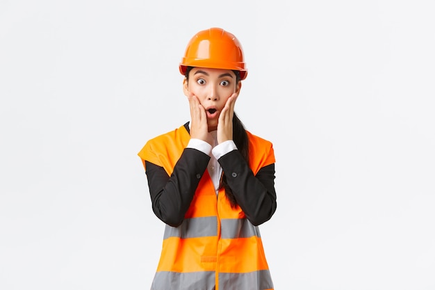 Zszokowana i zaniepokojona azjatycka inżynierka mająca problem w obszarze budowy, wpatruje się w projekt zdumiona z paniką na twarzy, trzymając ręce przy ustach i dysząc zmartwiona, stojąc na białym tle