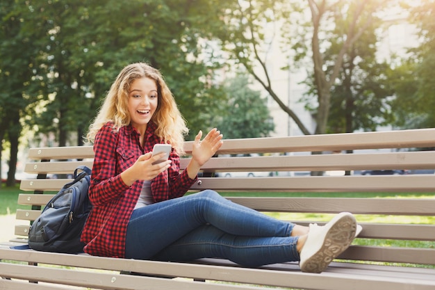 Zszokowana kobieta czytająca wiadomości na smartfonie na ławce na świeżym powietrzu. Koncepcja technologii, mediów społecznościowych, edukacji i freelancerów, kopia przestrzeń