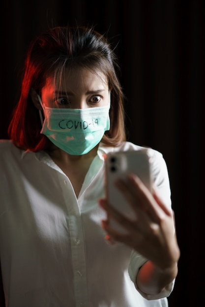 Zszokowana kobieta ubrana w maskę ochronną wirus koronowy (covid-19) używa telefonu, martwiąc się wiadomościami o wirusie koronowym.