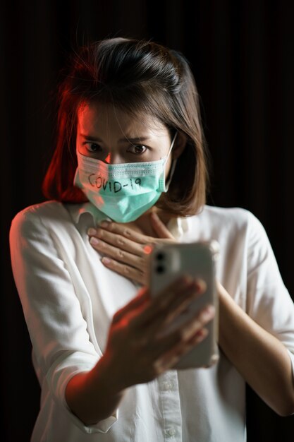 Zszokowana kobieta ubrana w maskę ochronną wirus koronowy używa smartfona, martwiąc się wiadomościami o wirusie koronowym.