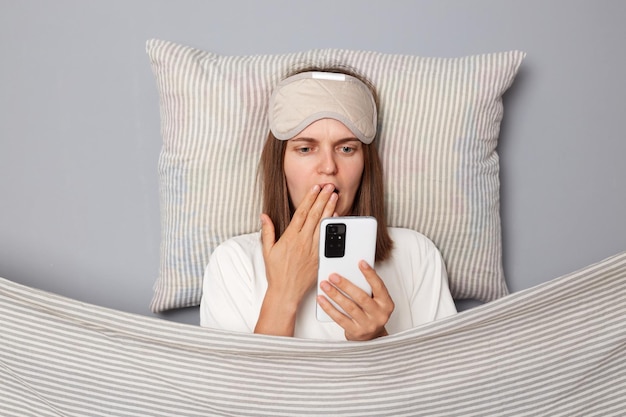 Zszokowana kobieta w białej koszulce i masce do spania leży w łóżku na poduszce pod kocem odizolowanym na szarym tle, trzymając telefon komórkowy zakrywający usta złe wieści