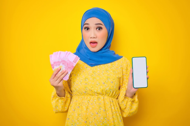 Zszokowana młoda Azjatycka kobieta w zwykłej sukience pokazująca pusty ekran telefon komórkowy i trzymająca banknoty na białym tle na żółtym tle