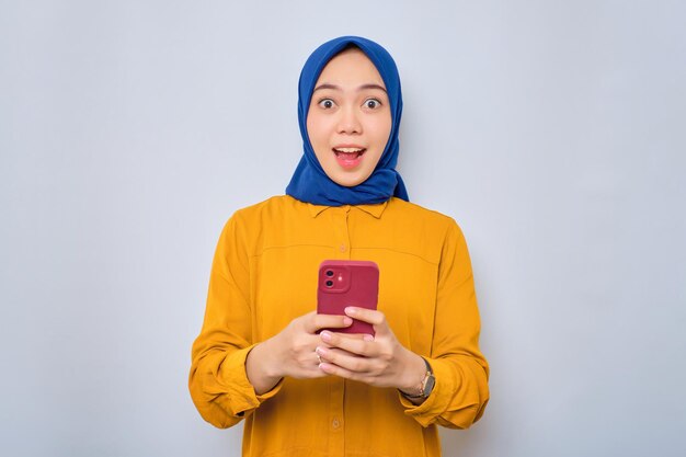 Zszokowana Młoda Azjatycka Muzułmanka Ubrana W Pomarańczową Koszulę, Trzymająca Telefon Komórkowy I Patrząca Na Kamerę Na Białym Tle Na Białym Tle
