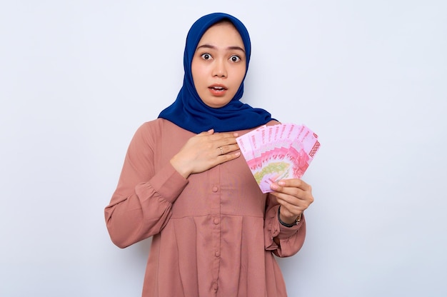 Zszokowana młoda azjatycka muzułmańska kobieta w różowej koszuli trzymająca banknoty pieniędzy na białym tle nad białym tle Koncepcja religijnego stylu życia ludzi