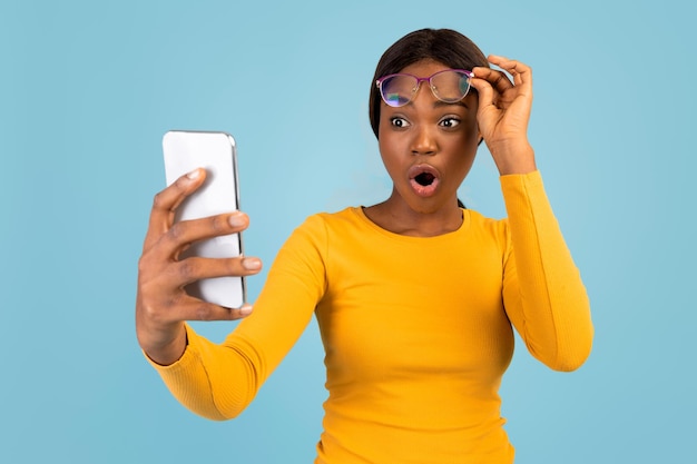 Zszokowana młoda czarna kobieta w żółtej koszulce zdejmuje okulary i patrzy na smartfona, czytając niesamowicie