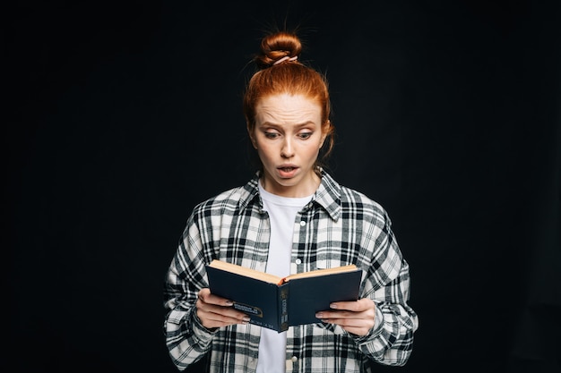 Zszokowana młoda kobieta studentka college'u czytająca książkę na czarnym tle na białym tle