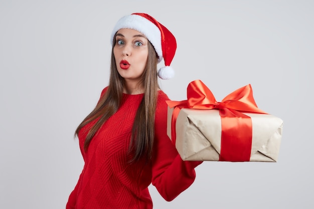 Zszokowana młoda kobieta w czerwonym swetrze, kobieta w czapce mikołaja, trzymając pudełko, na szarym tle. Koncepcja wakacje, Boże Narodzenie, Nowy Rok.