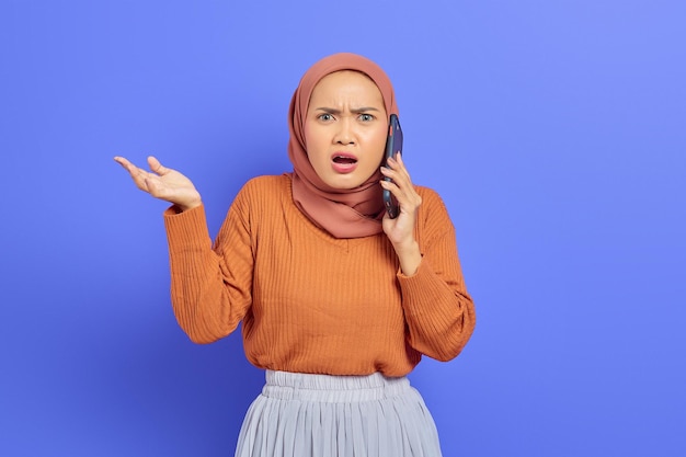 Zszokowana piękna Azjatycka kobieta w brązowym swetrze i hidżabie rozmawia przez telefon komórkowy z otwartymi ustami na białym tle na fioletowym tle. Koncepcja religijna ludzi islamu