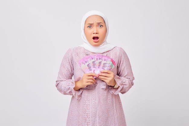 Zszokowana piękna młoda azjatycka muzułmanka ubrana w hidżab i fioletową sukienkę trzymająca banknoty rupii w gotówce na białym tle na tle białego studia