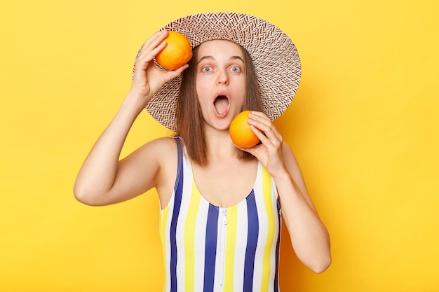 Zszokowana zdumiona kobieta ubrana w pasiasty kostium kąpielowy i kapelusz trzymająca pomarańcze patrząca na kamerę z zaskoczonym wyrazem twarzy odizolowana na żółtym tle