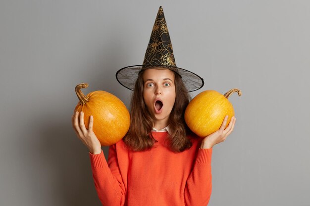 Zszokowana zdumiona kobieta w kapeluszu wiedźmy stojąca z dwiema pomarańczowymi dyniami w rękach na białym tle nad szarym tłem świętująca Halloween jest zdumiona patrząc na kamerę z otwartymi ustami