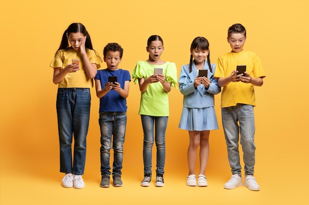 Zszokowane dzieci patrzące na ekrany telefonów komórkowych na żółtym tle