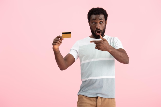 Zdjęcie zszokowany afroamerykanin wskazujący na kartę kredytową odizolowaną na różowo