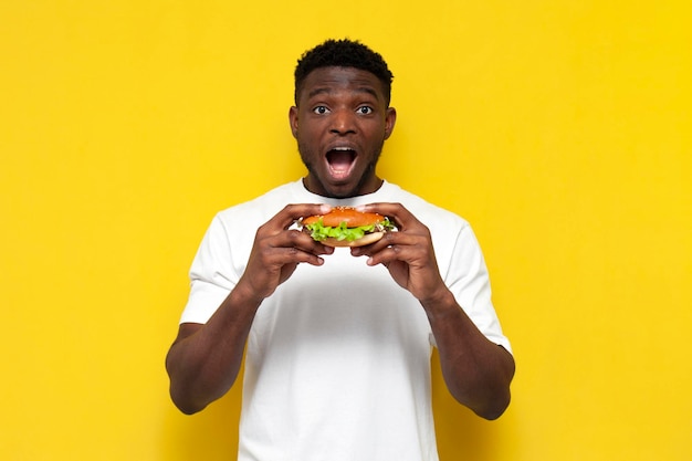 Zszokowany afrykański mężczyzna w białej koszulce trzymający dużego burgera i zaskoczony zdumiony facet jedzący fast foody