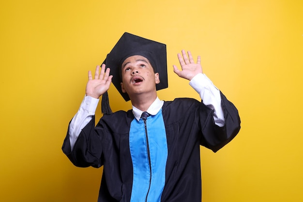 Zszokowany Azjat w ubraniach absolwentów patrzący na żółte tło