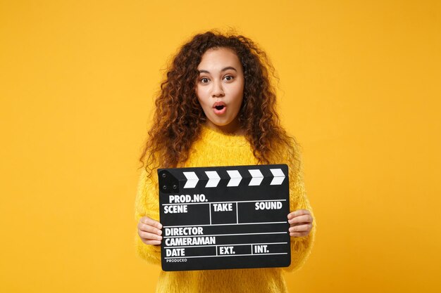 Zszokowany młoda african american dziewczyna w futro sweter pozowanie na białym tle na żółtym pomarańczowym tle w studio. Koncepcja życia ludzi. Makieta miejsca na kopię. Trzymając klasyczny czarny film clapperboard.