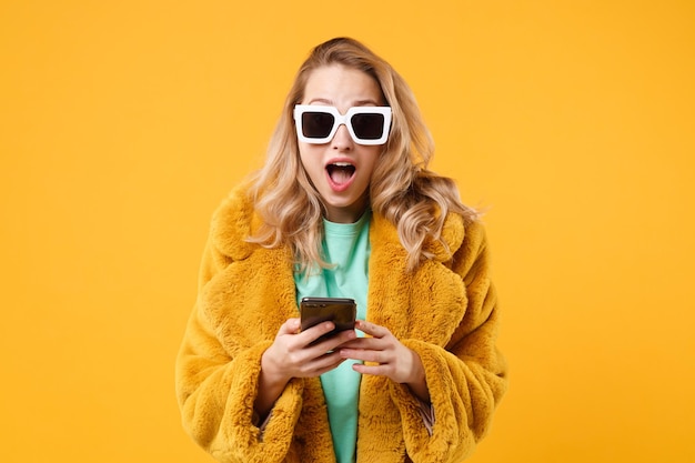 Zszokowany młoda blondynka kobieta dziewczyna w żółtym futrze, ciemne okulary pozowanie na białym tle na pomarańczowym tle w studio. Koncepcja życia ludzi. Makieta miejsca na kopię. Za pomocą telefonu komórkowego wpisując wiadomość sms.