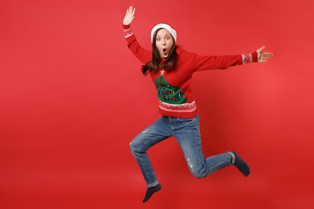 Zszokowany młoda dziewczyna Santa trzymając szeroko otwarte usta, patrząc zaskoczony skoki rozłożone ręce, nogi na białym tle na czerwonym tle. Szczęśliwego nowego roku 2019 celebracja party wakacje koncepcja. Makieta miejsca na kopię.